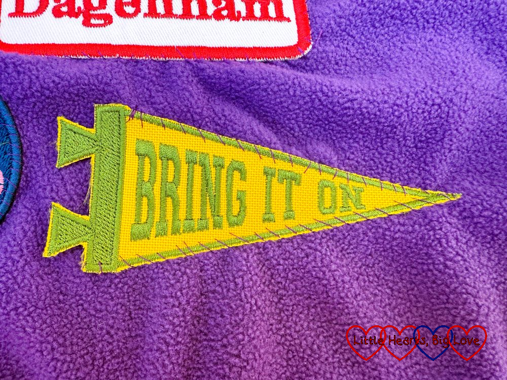 A yellow pennant shaped sew-on patch with 'Bring It On' in green writing on a purple blanket