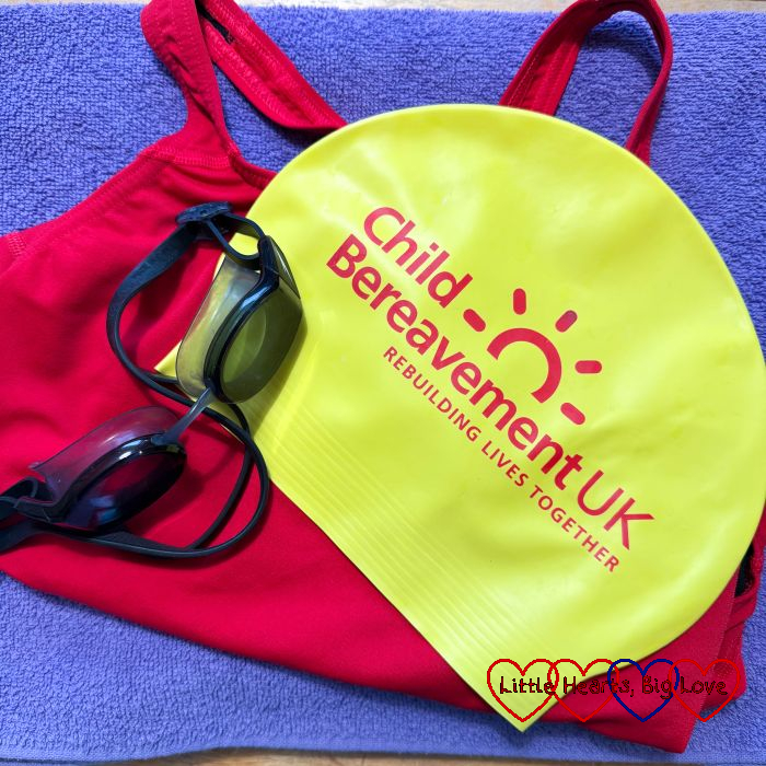 A yellow "Child Bereavement UK" swim hat with black googles on a red swimsuit on top of a purple towel