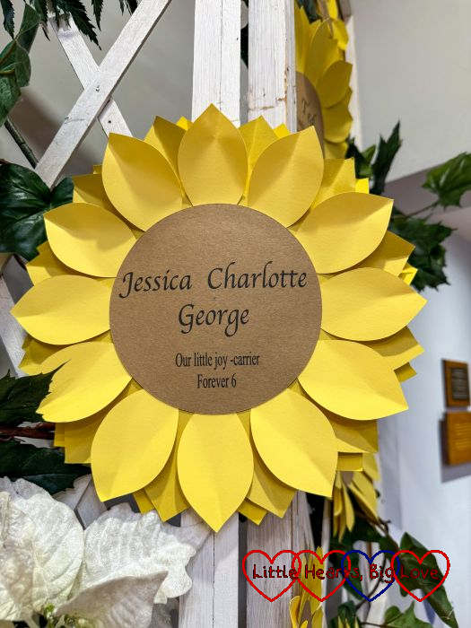 A paper sunflower on a trellis with the words "Jessica Charlotte George, our little joy-carrier. Forever 6"