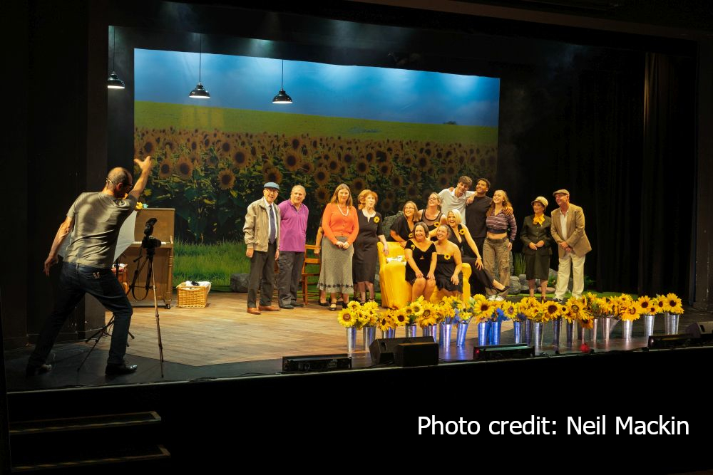 A scene from Calendar Girls showing a photographer taking a photo of two ladies in black dresses sitting on a yellow sofa with a group gathered around them