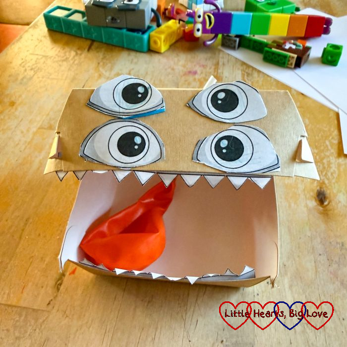 A monster with four eyes made from a cardboard box with a balloon tongue