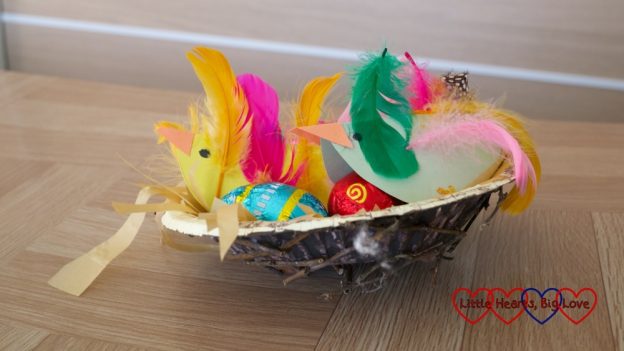 A paper bowl nest with twigs glued to it and paper birds inside covered in feathers looking after small chocolate eggs