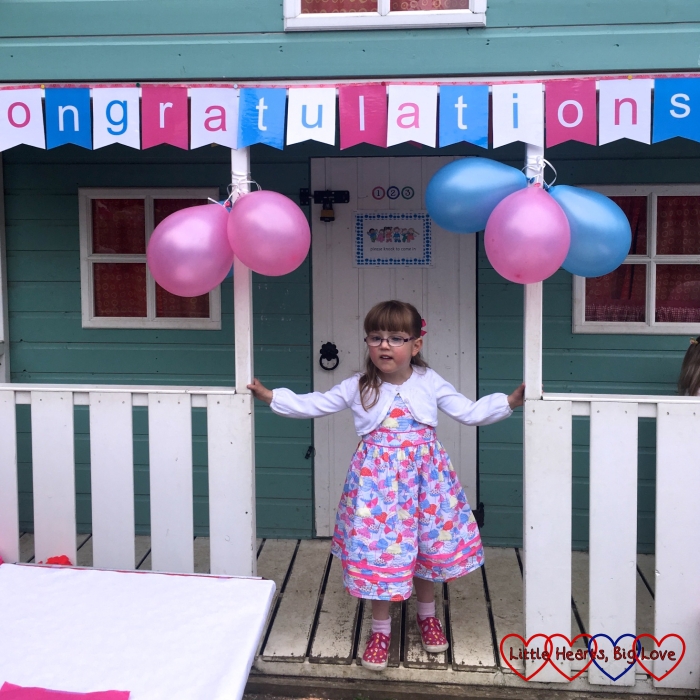 Jessica standing in front of the playhouse at preschool with a banner saying "Congratulations" over her head at her preschool leavers' party