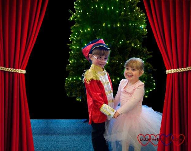 Jessica dressed in a soldier costume and Sophie in a pink tutu standing in front of a Christmas tree with stage curtains either side of them