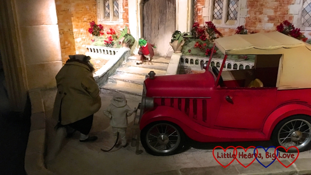 Toad on the steps of Toad Hall with his motor car outside