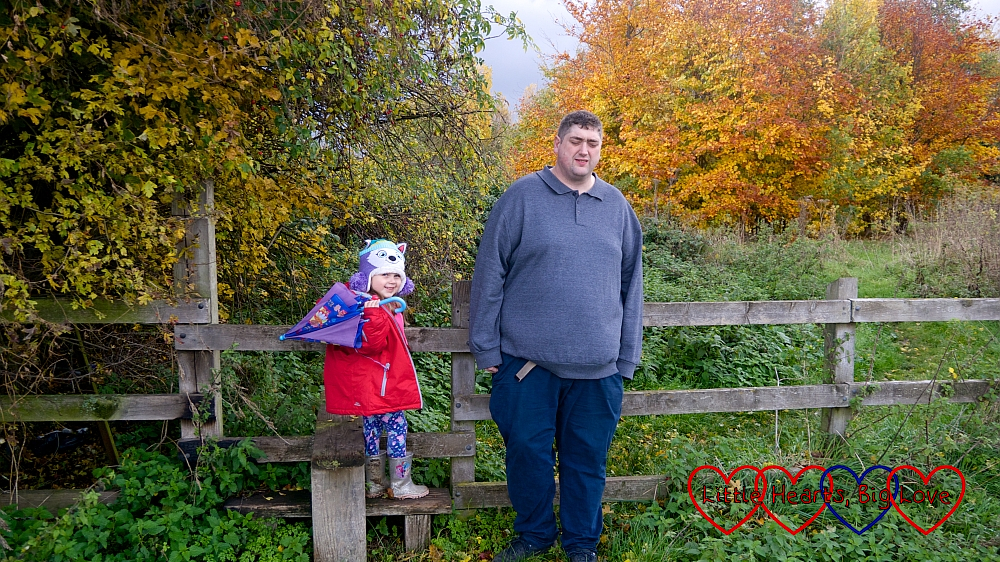 Sophie standing on a stile next to hubby in front of some autumn trees