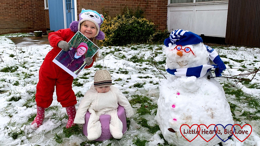 Sophie (holding a picture of Jessica) in the snow with Thomas in the Bumbo and a snowman with pipe-cleaner glasses and a blue and white hat and scarf