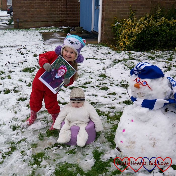 Sophie (holding a picture of Jessica) in the snow with Thomas in the Bumbo and a snowman with pipe-cleaner glasses and a blue and white hat and scarf