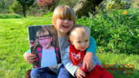 Sophie and Thomas sitting in Grandma's garden, with Sophie holding the picture of Jessica