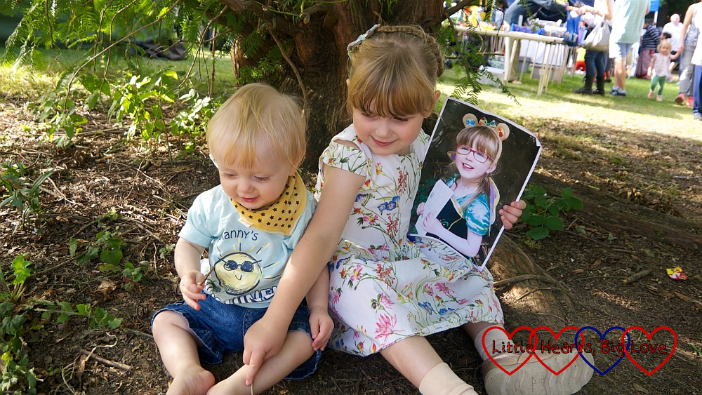 Sophie and Thomas sitting under a tree at Pinner Village Show. Sophie is holding a picture of Jessica