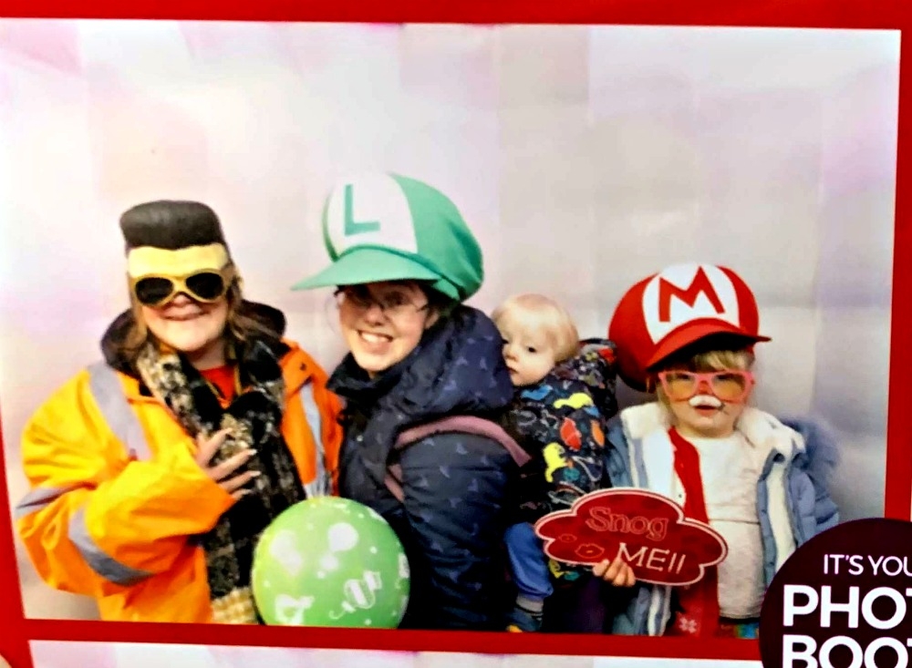 My sister wearing an Elvis wig and glasses, me wearing a Luigi hat with Thomas in the carrier on my back and Sophie with her face painted like a reindeer wearing a Mario hat and glasses in the festive photo booth