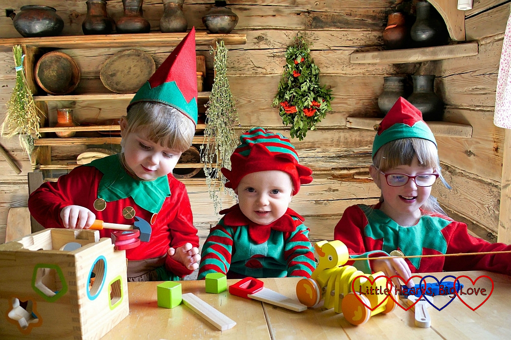 Sophie, Thomas and Jessica dressed as Christmas elves at the table together with a log cabin background