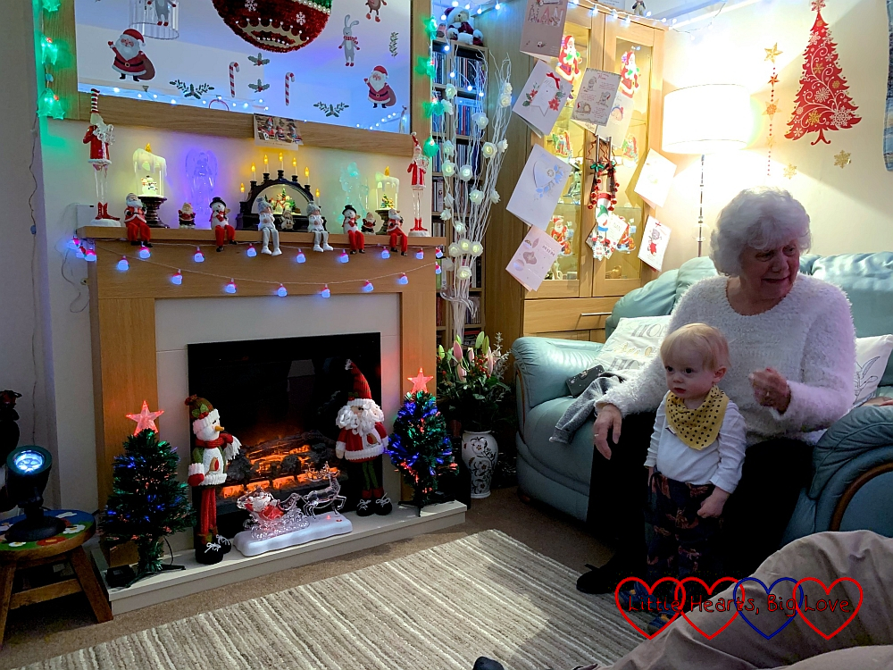 My mum's living room decorated like a Christmas grotto with my mum sitting on the sofa and Thomas standing in front of her looking at the decorations