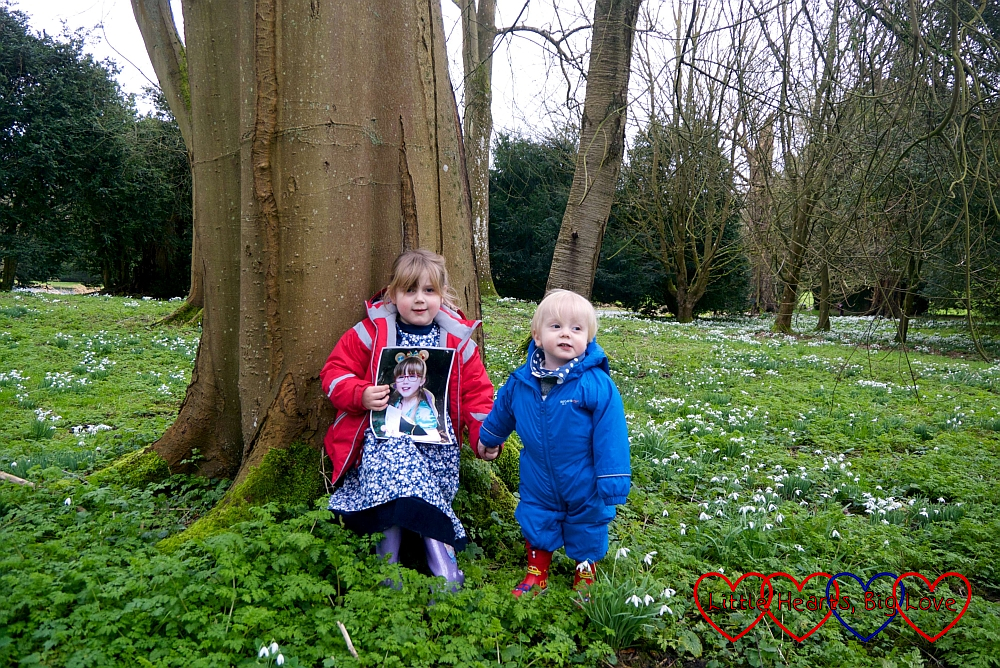 Sophie standing leaning against a tree, holding a photo of Jessica in front of her, and Thomas's hand as he stands next to her with a field of snowdrops surrounding them