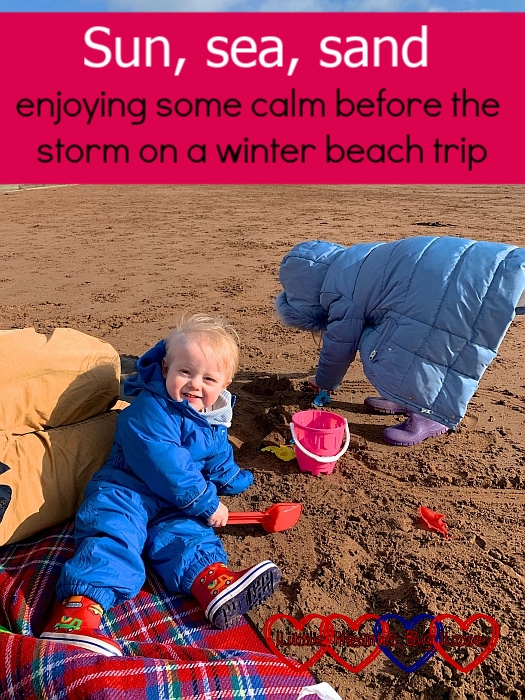 A smiley Thomas sitting on a picnic blanket on the beach holding a spade, while Sophie digs in the sand behind him - "Sun, sea and sand - enjoying some calm before the storm on a winter beach trip"