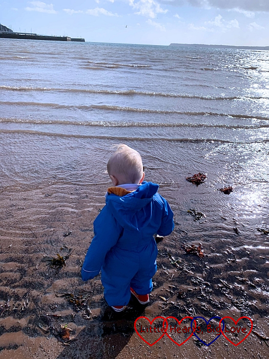 Thomas in his blue puddlesuit and red wellies, standing on the edge of the water at the beach in Torquay