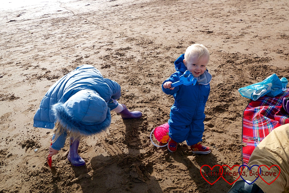 A smiley Thomas holding a rake while Sophie digs in the sand next to him