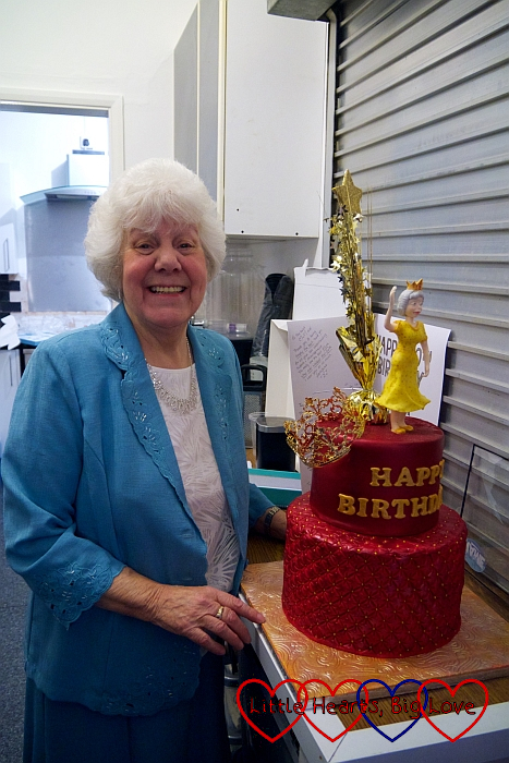 My mum with her 80th birthday cake (a two tier cake with red icing and a modelled figure of a queen on the top)