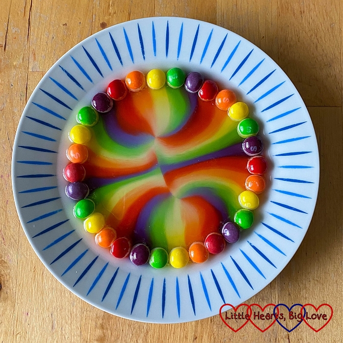 Skittles around the edge of a plate with rainbow patterns in the middle of the plate