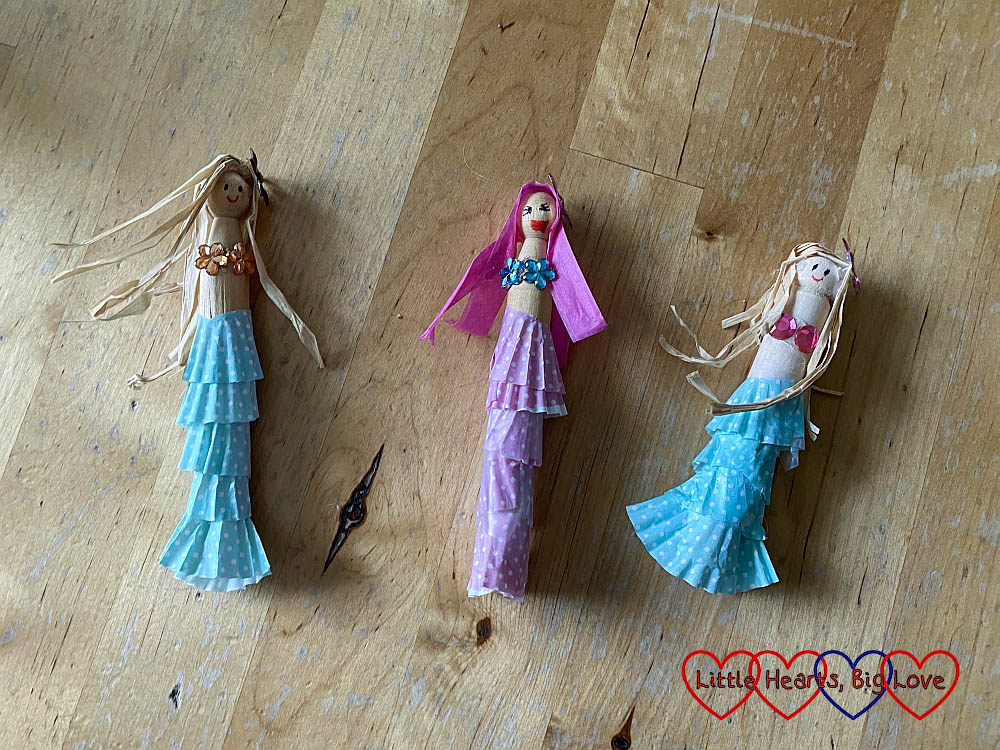 Three mermaid peg dolls with petits-fours cases tails, raffia hair and gems stuck on for a bikini top. Two have blue tails, and beige raffia hair; one has a pink tail and pink raffia hair