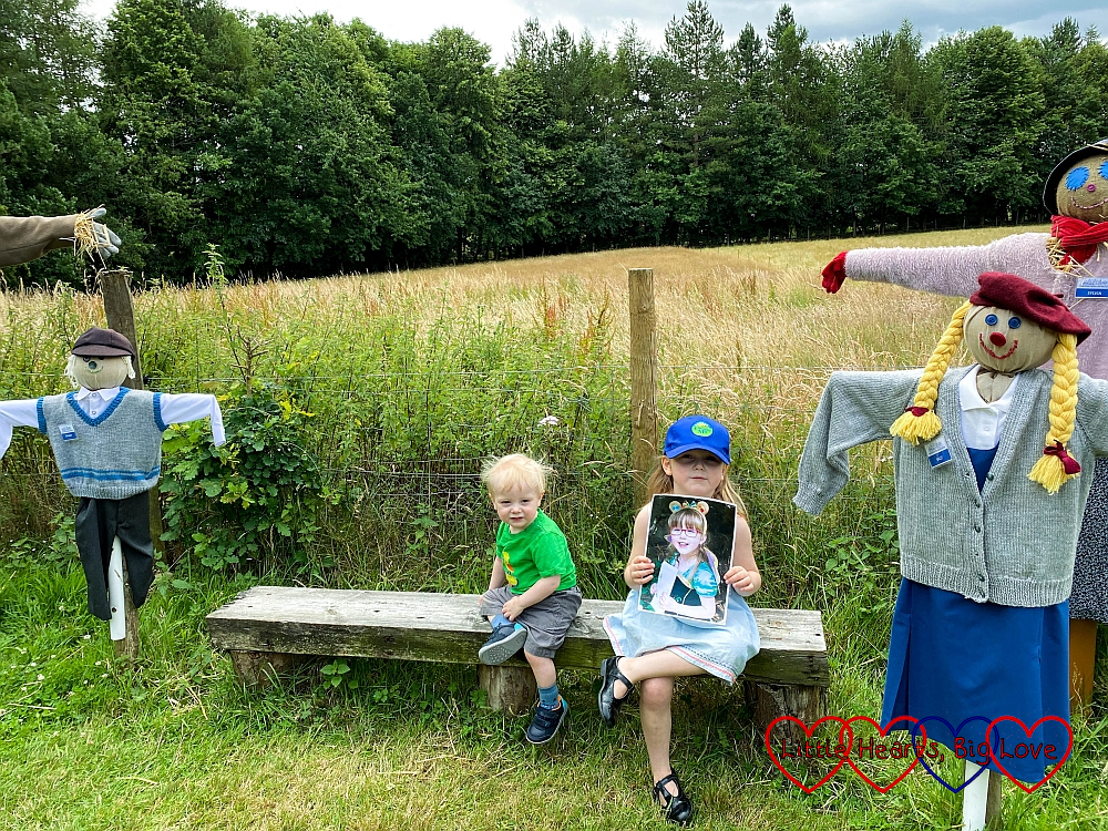 Sophie (holding a photo of Jessica) and Thomas sitting on a bench in between a family of scarecrows