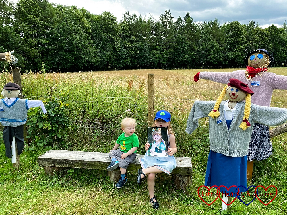 Sophie (holding a photo of Jessica) and Thomas sitting on a bench in between a family of scarecrows