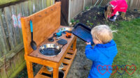 Thomas standing by the mud kitchen holding a tray