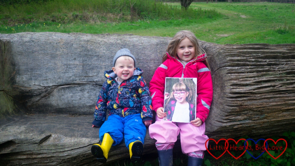 Sophie and Thomas sitting on a seat carved in a fallen tree with Sophie holding a picture of Jessica