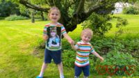 Sophie and Thomas wearing matching rainbow T-shirts and blue shorts standing in Grandma and Grandad's garden with Sophie holding a photo of Jessica