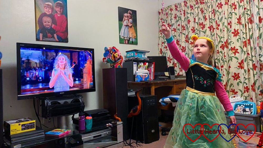 Sophie posing in a princess dress and holding a wand aloft with Strictly on the TV behind her