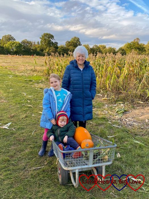 Sophie standing next to Nanny in front of a corn field with Thomas sitting in a trolley in front with two pumpkins and some corn cobs next to him