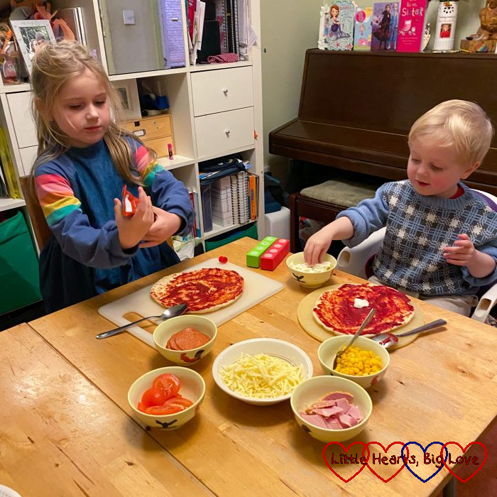 Sophie and Thomas with pizza bases covered in tomato puree, choosing toppings from a selection in bowls on the table