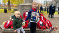 Sophie (holding a photo of Jessica on Remembrance Sunday) and Thomas standing in front of poppy wreaths on the peace memorial