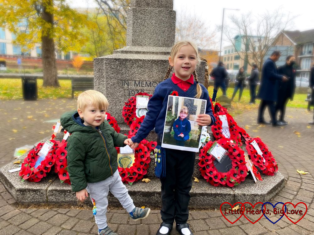 Sophie (holding a photo of Jessica on Remembrance Sunday) and Thomas standing in front of poppy wreaths on the peace memorial