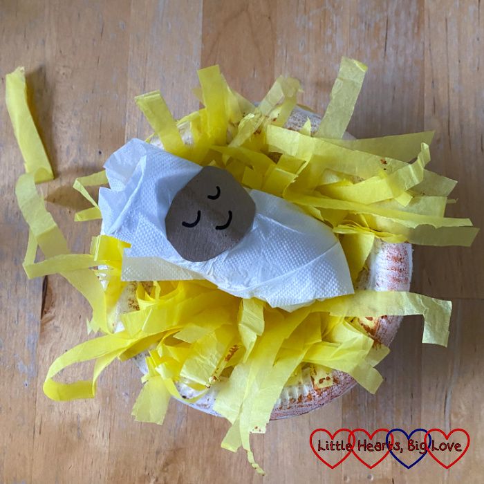A cardboard tube baby Jesus in a paper bowl manger filled with shredded yellow tissue paper