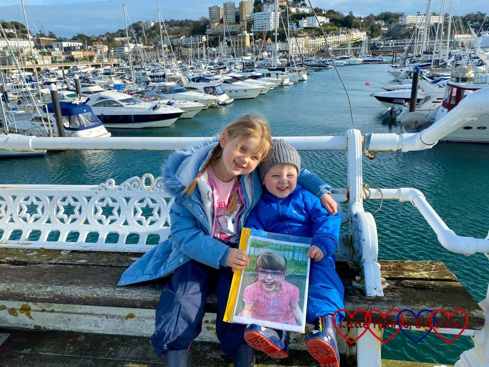 Sophie and Thomas sitting on a bench on the pier at Torquay with boats on the water behind them and them both holding a photo of Jessica