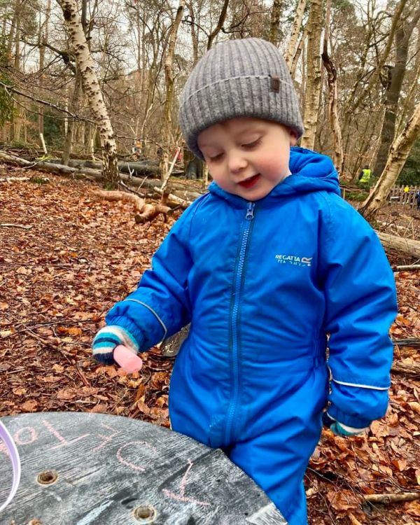 Thomas holding a piece of chalk having written 'Tot' on a cable drum at forest school