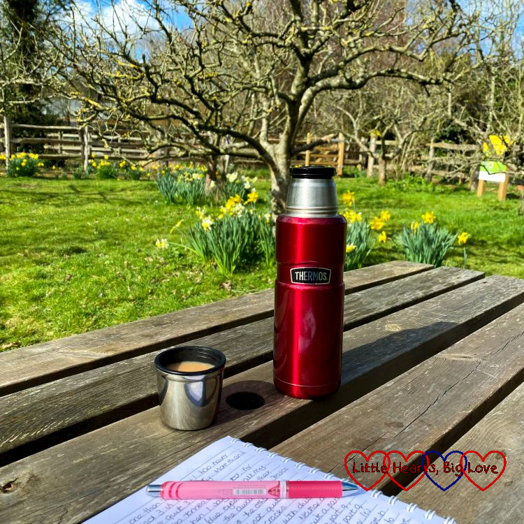 A notebook, pen and flask of tea on a wooden picnic table looking over an apple tree and some daffodils