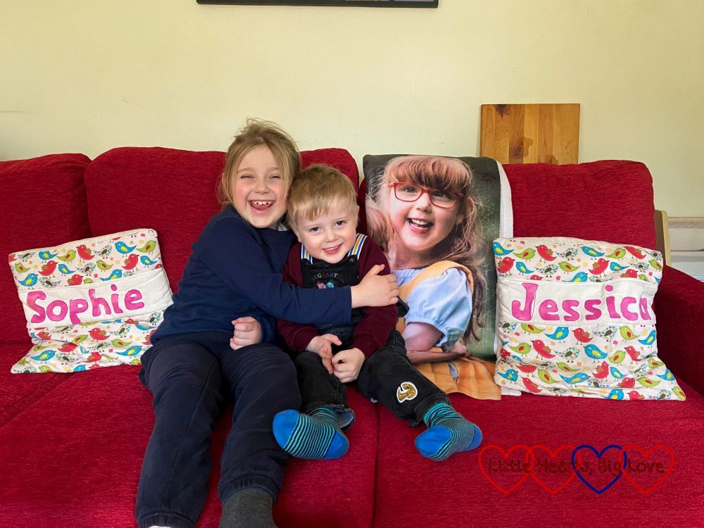 Sophie and Thomas sitting on the sofa with an image of Jessica from her photo blanket draped on the sofa behind them. There is a cushion that says 'Sophie' next to Sophie and another one that says 'Jessica' on the opposite side, next to the photo of Jessica