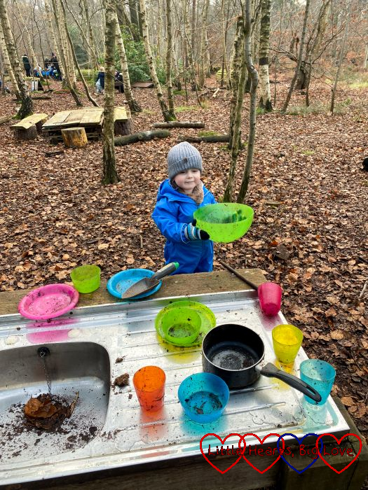 Thomas in a puddle suit, scarf and hat playing with the mud kitchen at forest school