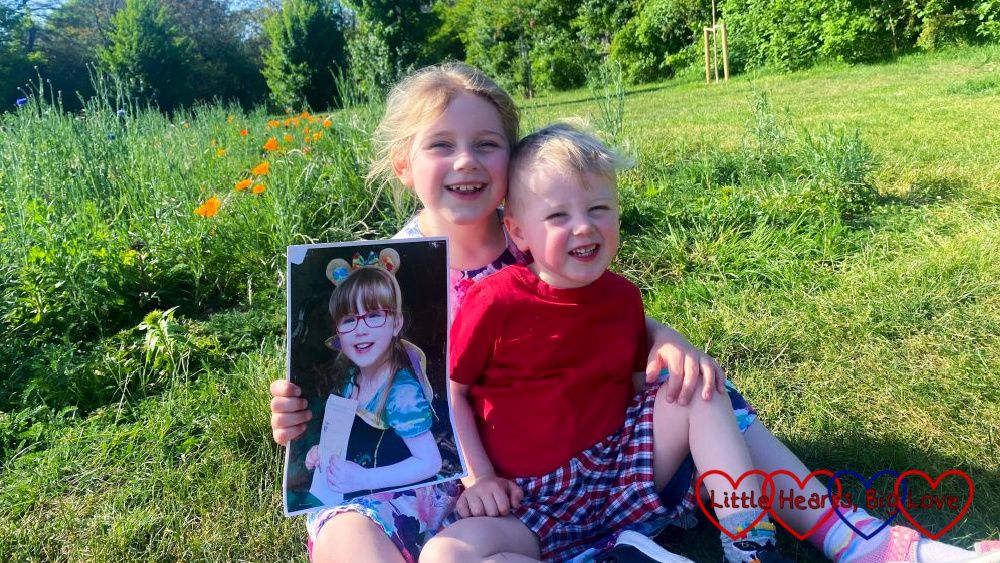Sophie and Thomas sitting on the grass in front of a wildflower patch with Sophie holding a photo of Jessica