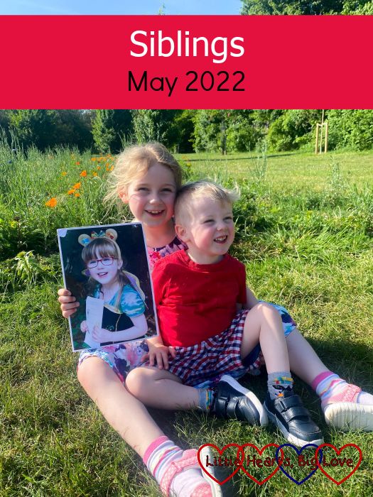 Sophie and Thomas sitting on the grass in front of a wildflower patch with Sophie holding a photo of Jessica - "Siblings - May 2022"
