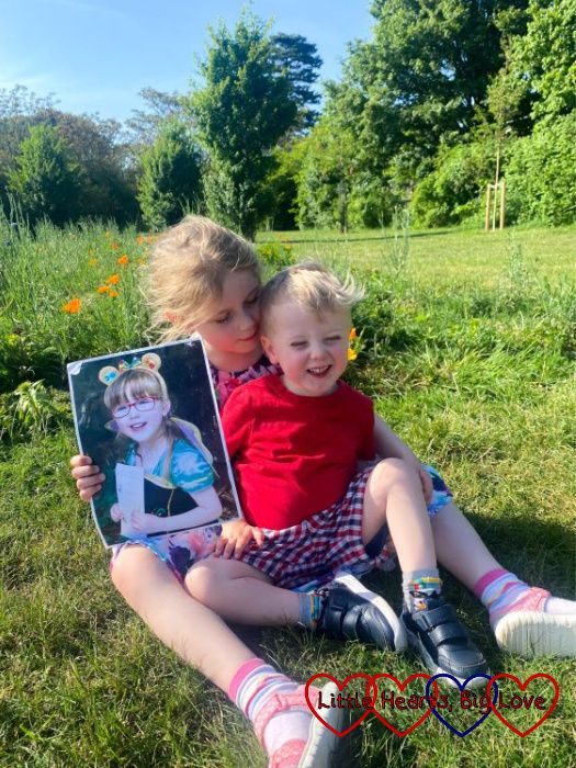 Sophie and Thomas sitting on the grass in front of a wildflower patch with Sophie holding a photo of Jessica