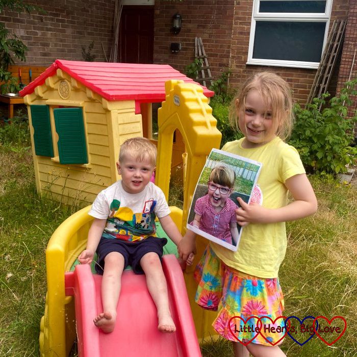 Sophie standing next to the little slide in the garden holding a photo of Jessica and Thomas sitting at the top of the slide