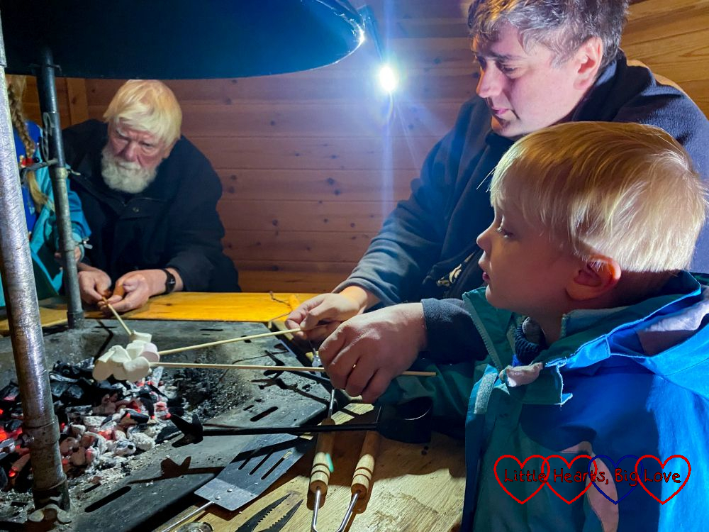 Thomas toasting marshmallows over a fire with help from Daddy while Grandad watches