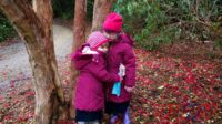 Sophie and Jessica wearing winter coats and hats, standing in front of a tree hugging each other