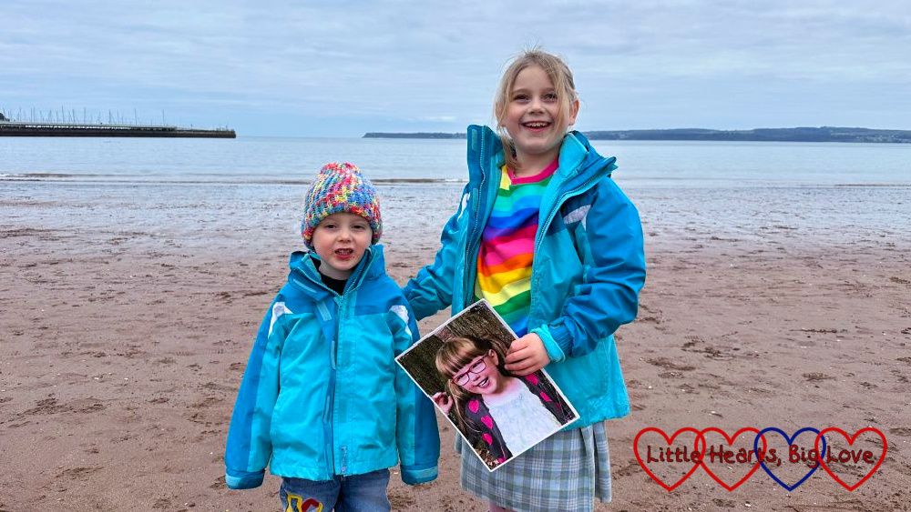 Sophie and Thomas standing on the beach at Torquay in matching blue coats. Sophie is holding a picture of Jessica.