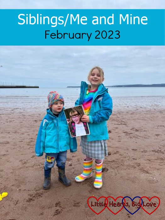 Sophie and Thomas standing on the beach at Torquay in matching blue coats. Sophie is holding a picture of Jessica. - "Siblings/Me and Mine - February 2023"