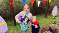 Sophie (holding an Easter egg and a photo of Jessica) and Thomas standing near cardboard cut out eggs