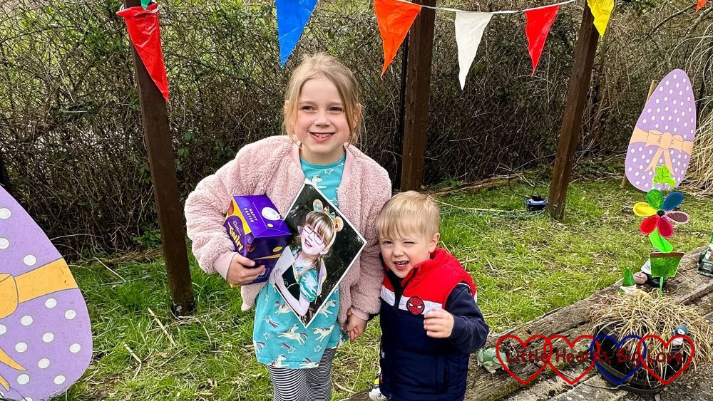 Sophie (holding an Easter egg and a photo of Jessica) and Thomas standing near cardboard cut out eggs